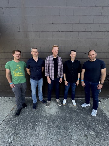 The five members of The Whiles standing together in front of a dark brick wall